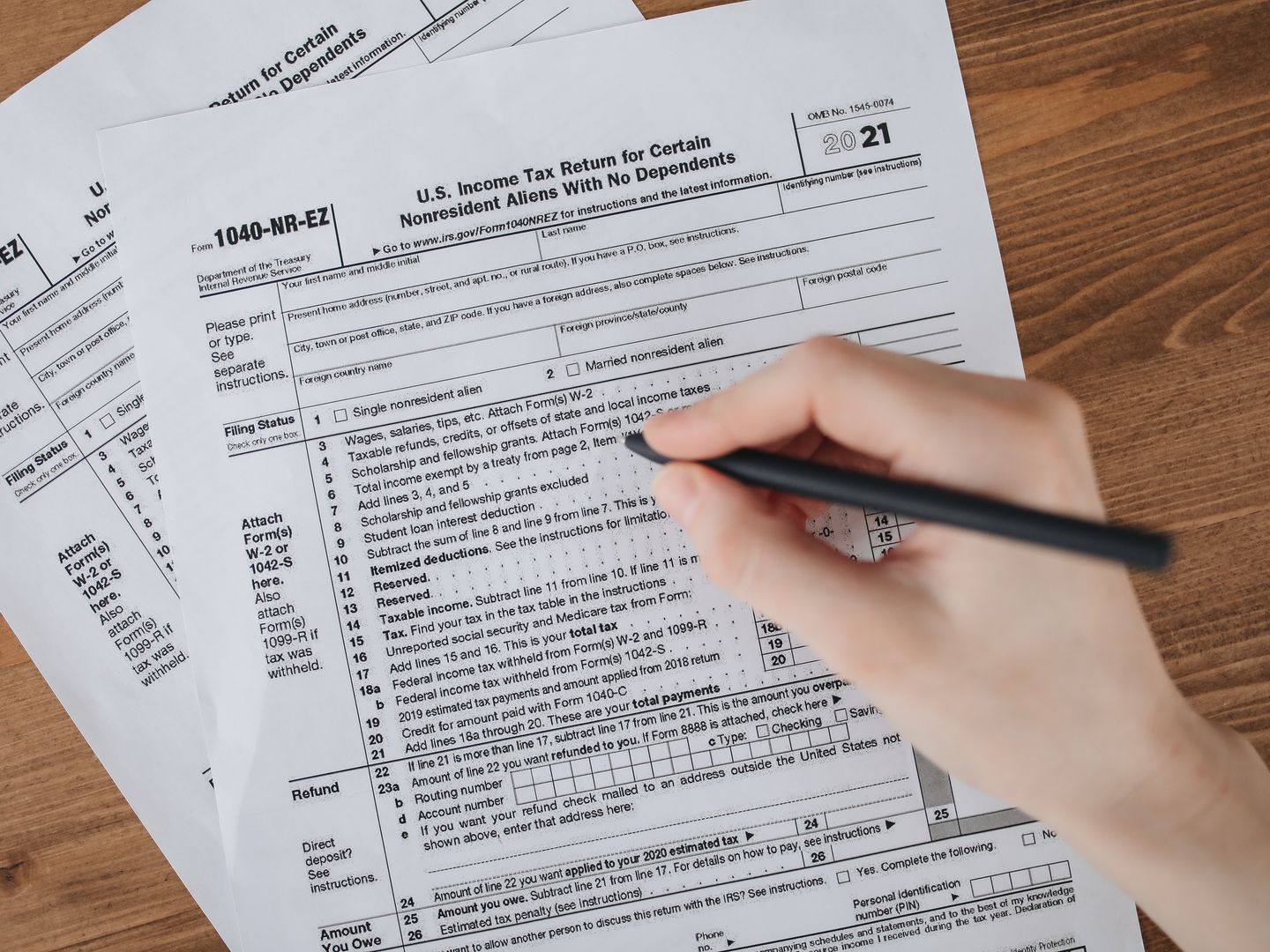 Person filling out U.S. tax form 1040 NR EZ with a pen on a wooden table.