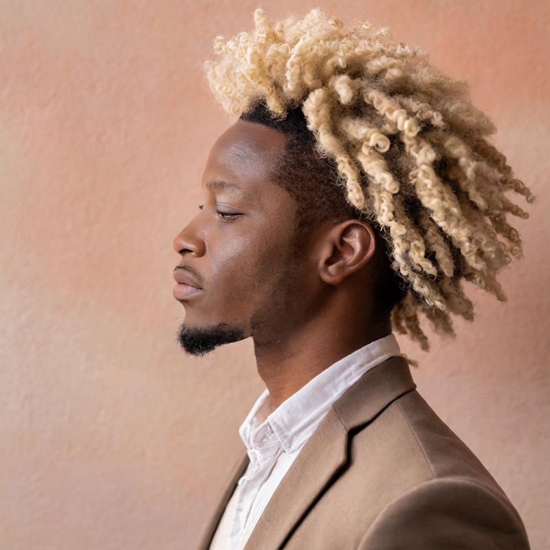 Profile portrait of a stylish man with blonde curly hair in a beige blazer against a textured background.